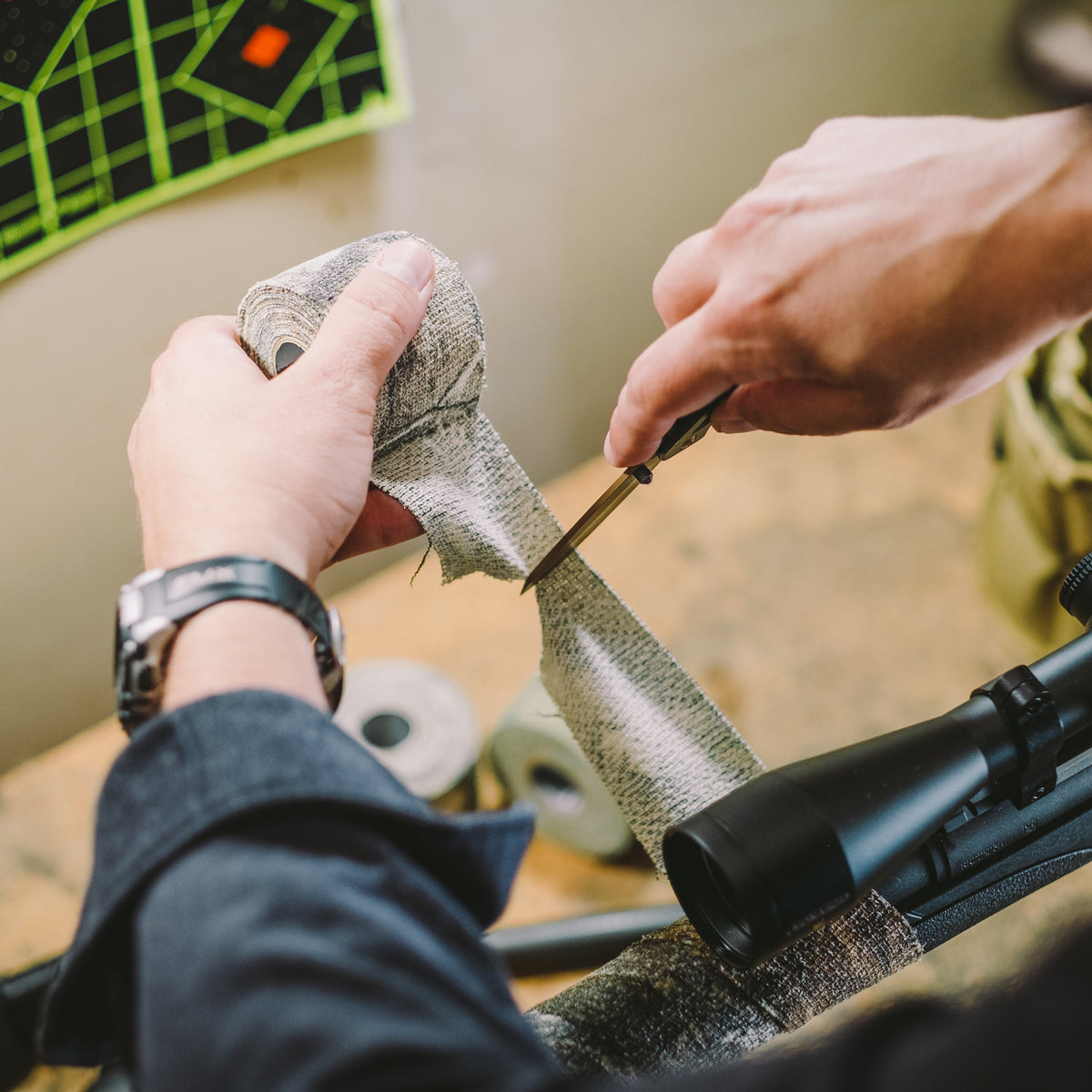 Hands cutting a strip of Camo Form with a knife beside a scoped rifle