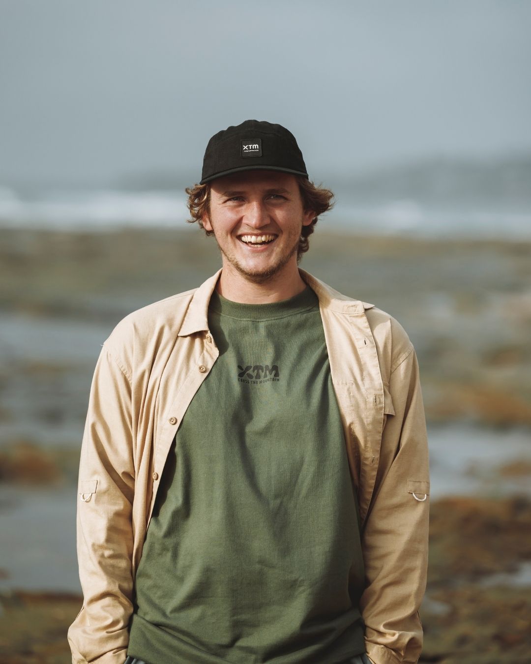 Smiling man in a tan jacket and green tee wearing the XTM Hakea cap in Black by the water