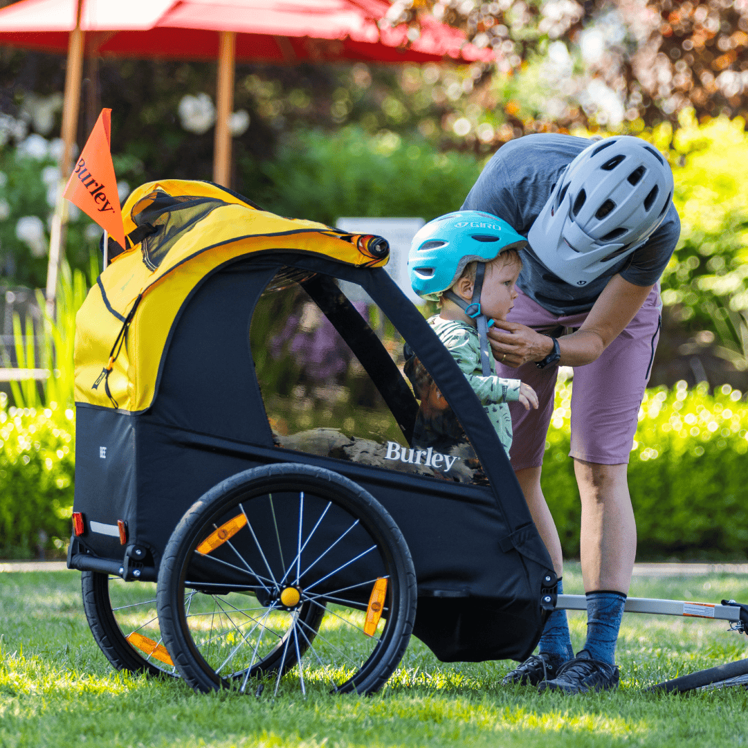 Parent buckling a child in a helmet into a Burley Bee trailer on a lawn