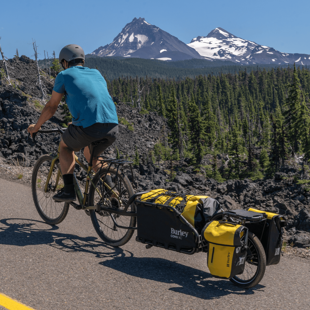 Rider on a paved road towing a loaded Burley Coho XC with snow-capped peaks behind