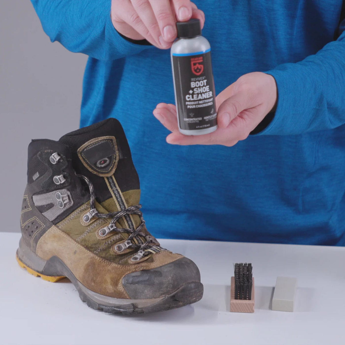 Hand holding the cleaner bottle above a muddy leather hiking boot, brush and eraser block nearby