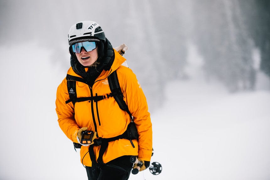 Skier in an orange jacket and white helmet in falling snow wearing a transceiver harness