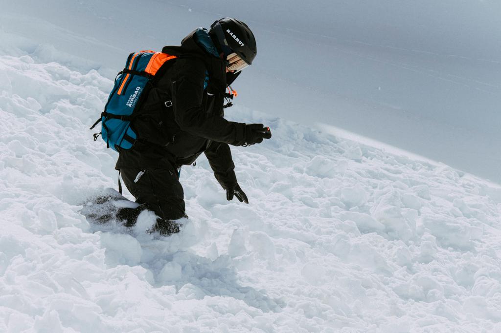 Skier in black kneeling on avalanche debris during a transceiver search, blue pack on their back