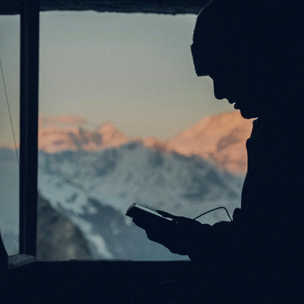 Silhouette of a person at a hut window holding an avalanche transceiver at sunrise
