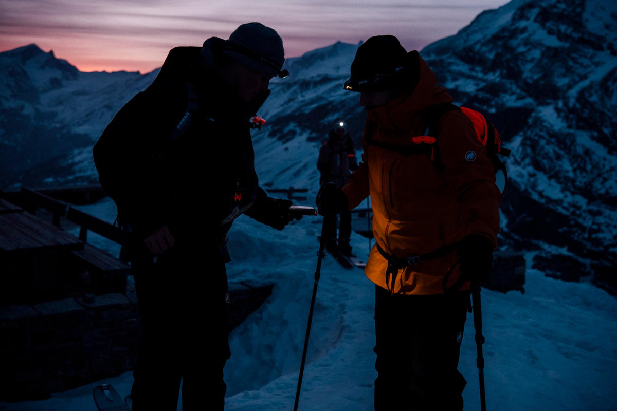 Two ski tourers checking their avalanche beacons by head torch at dusk with mountains behind