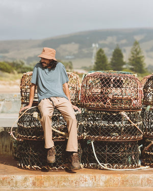 Man in the Woodsmoke XTM Sundew bucket hat sitting on stacked fishing pots by the coast