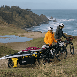 Two riders on a coastal headland, Burley Coho XC loaded with bags and a surfboard behind a bike