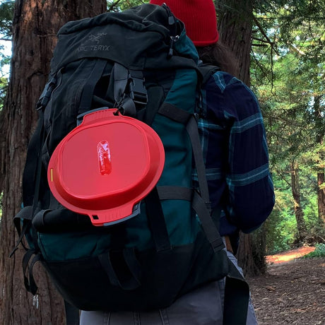 Closed red and charcoal humangear GoKit clipped to the outside of a green hiking backpack on a forest trail