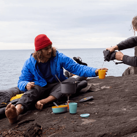 Hiker on a rocky shoreline holding a yellow Passage cup, teal bowl and cup by the stove
