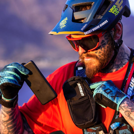 Mountain biker in a helmet and goggles pulling a phone from a USWE pocket on his chest